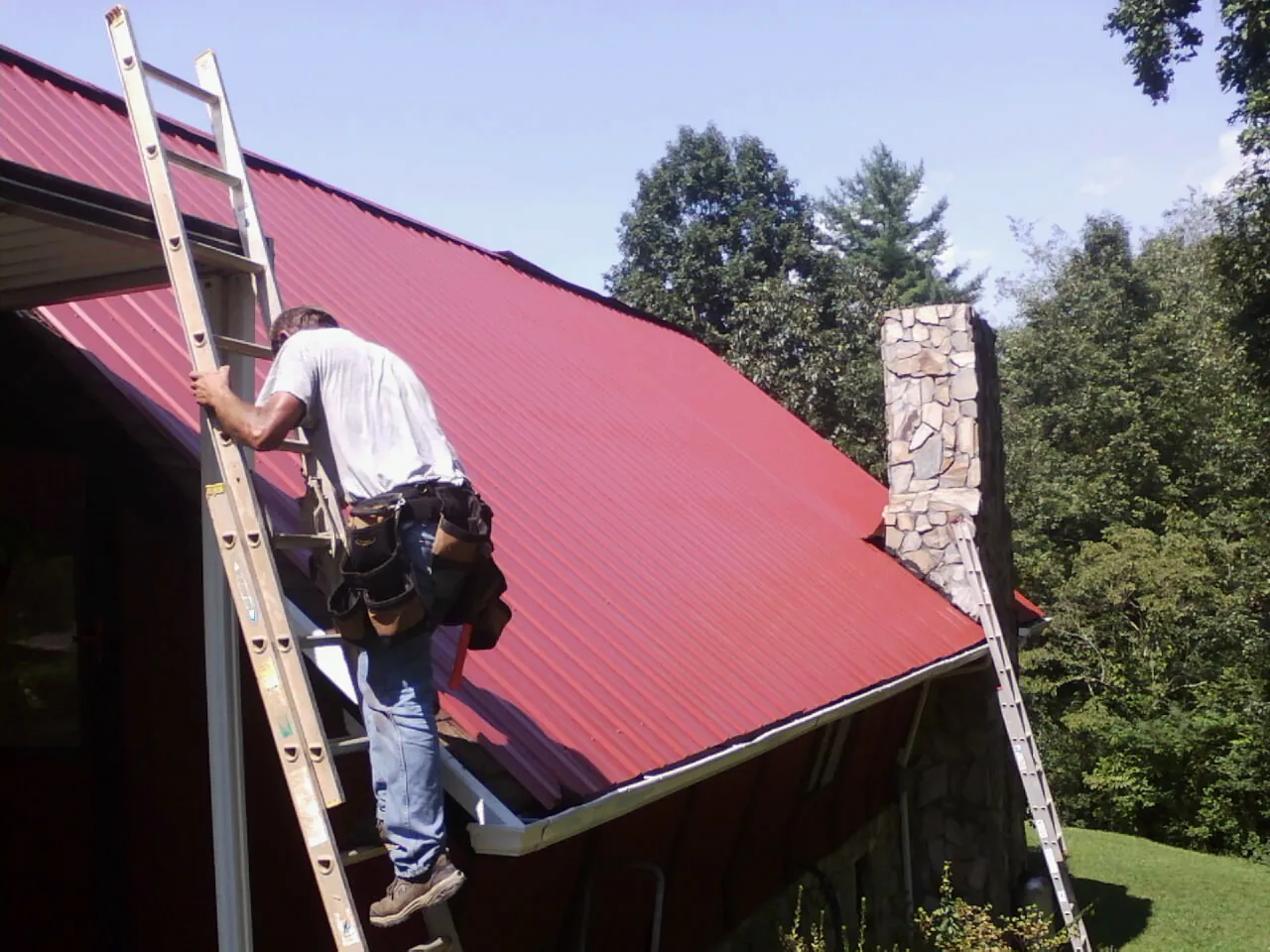 Roofer on a ladder beside a red metal roof and stone chimney — Soffit & Fascia Repair by Roofing Brothers in Concord